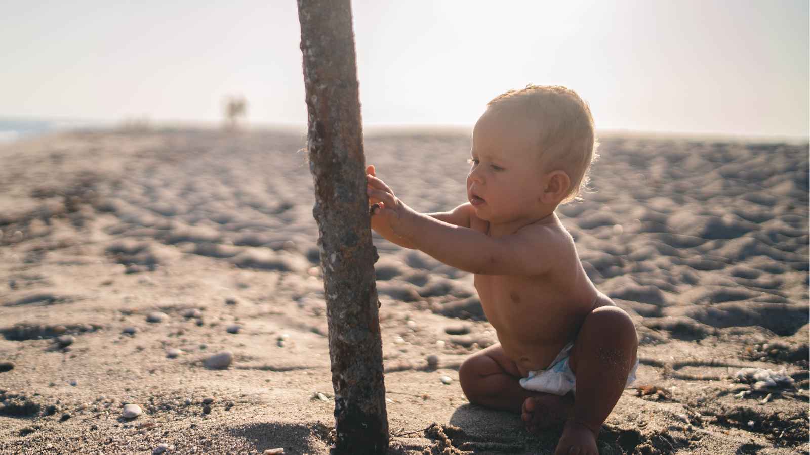 Baby on sandy beach touching a wooden pole with a curious expression.