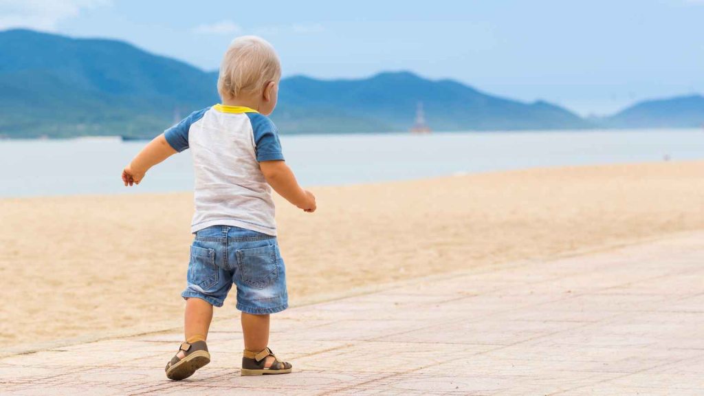 A toddler in a white and blue shirt and denim shorts walks on a beachside path with mountains in the background.