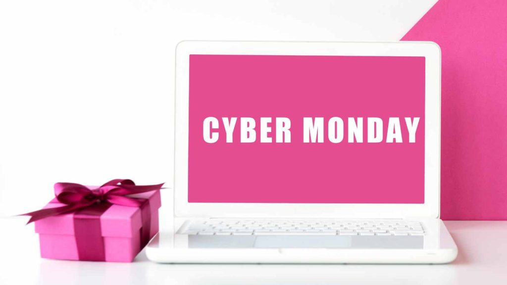 Open laptop displaying "Cyber Monday" in white text on a pink screen. Nearby, a small pink gift box with a bow.