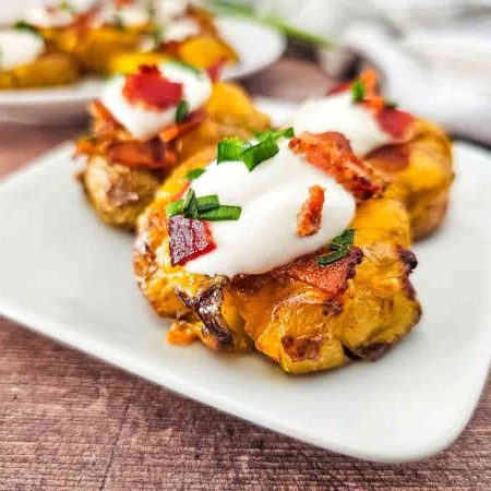 A plate of loaded smashed potatoes topped with sour cream, bacon bits, and chopped green chives.