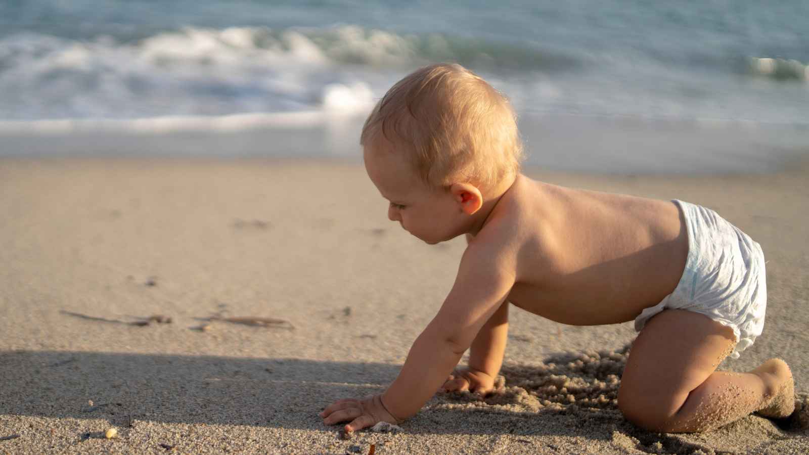 A baby in a diaper crawls on a sandy beach near the ocean.