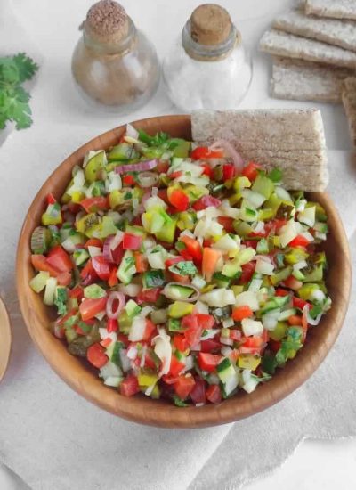 A wooden bowl filled with pickle de gallo salsa, surrounded by whole grain crackers and wooden utensils, on a white cloth.