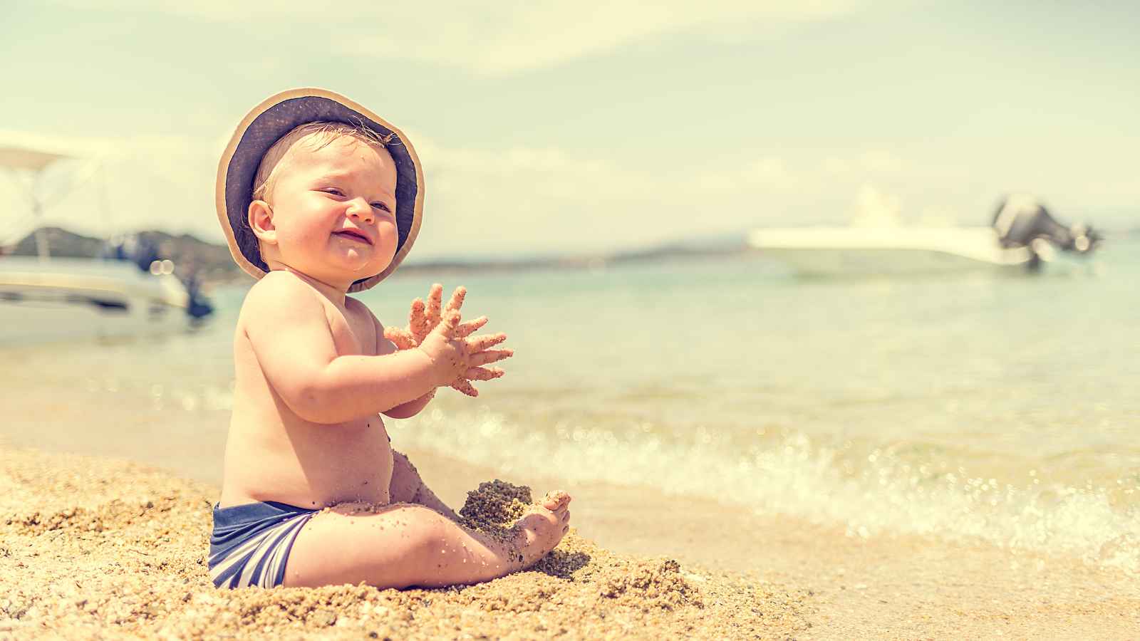 A baby wearing a hat sits on a sandy beach, playing with sand. A boat is visible in the background on the water.