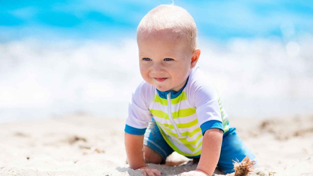 A toddler in a striped shirt crawls on a sandy beach with the ocean in the background.