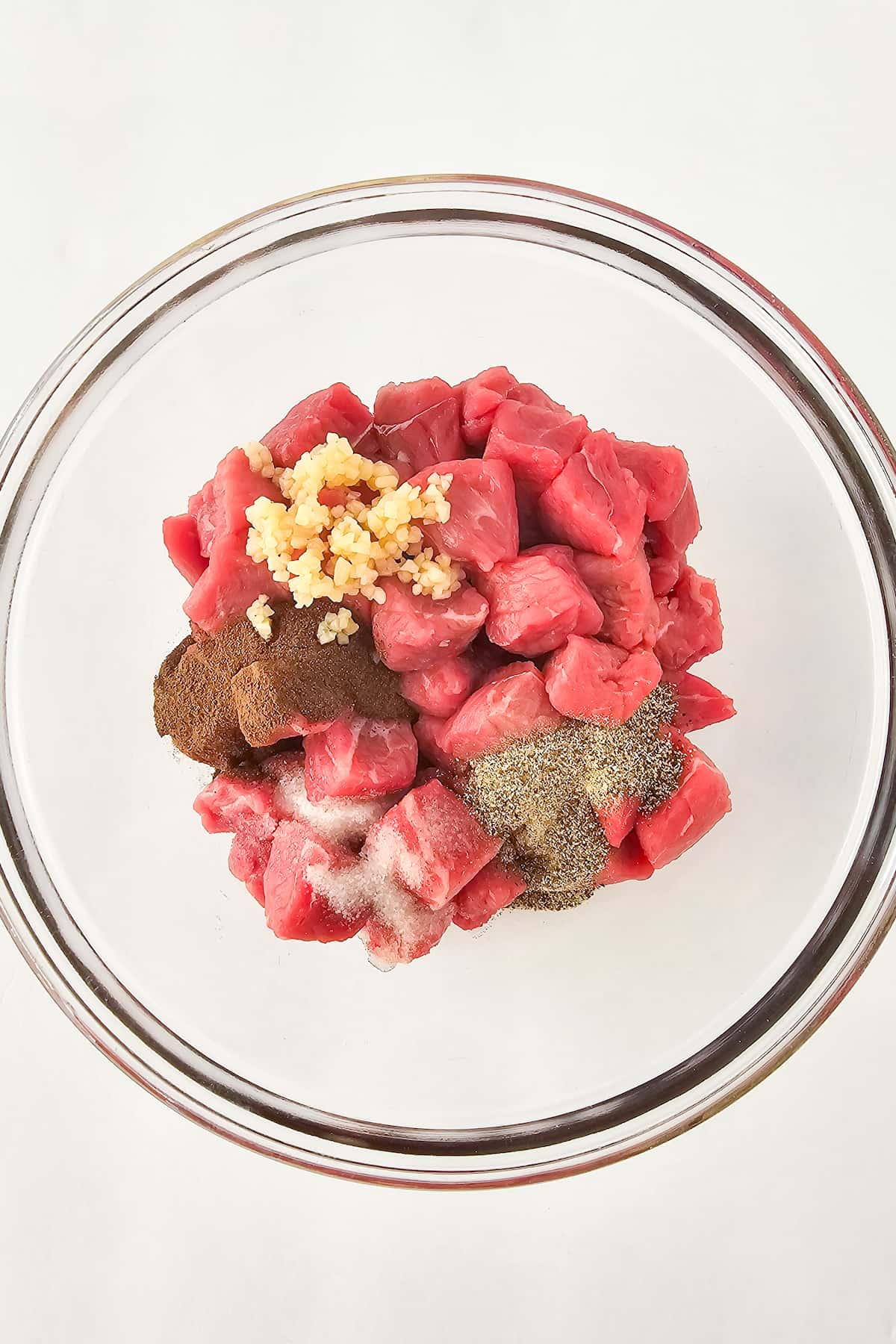 A glass bowl with raw, cubed beef prepared for air fryer steak bites, minced garlic, and various spices all set against a white background.