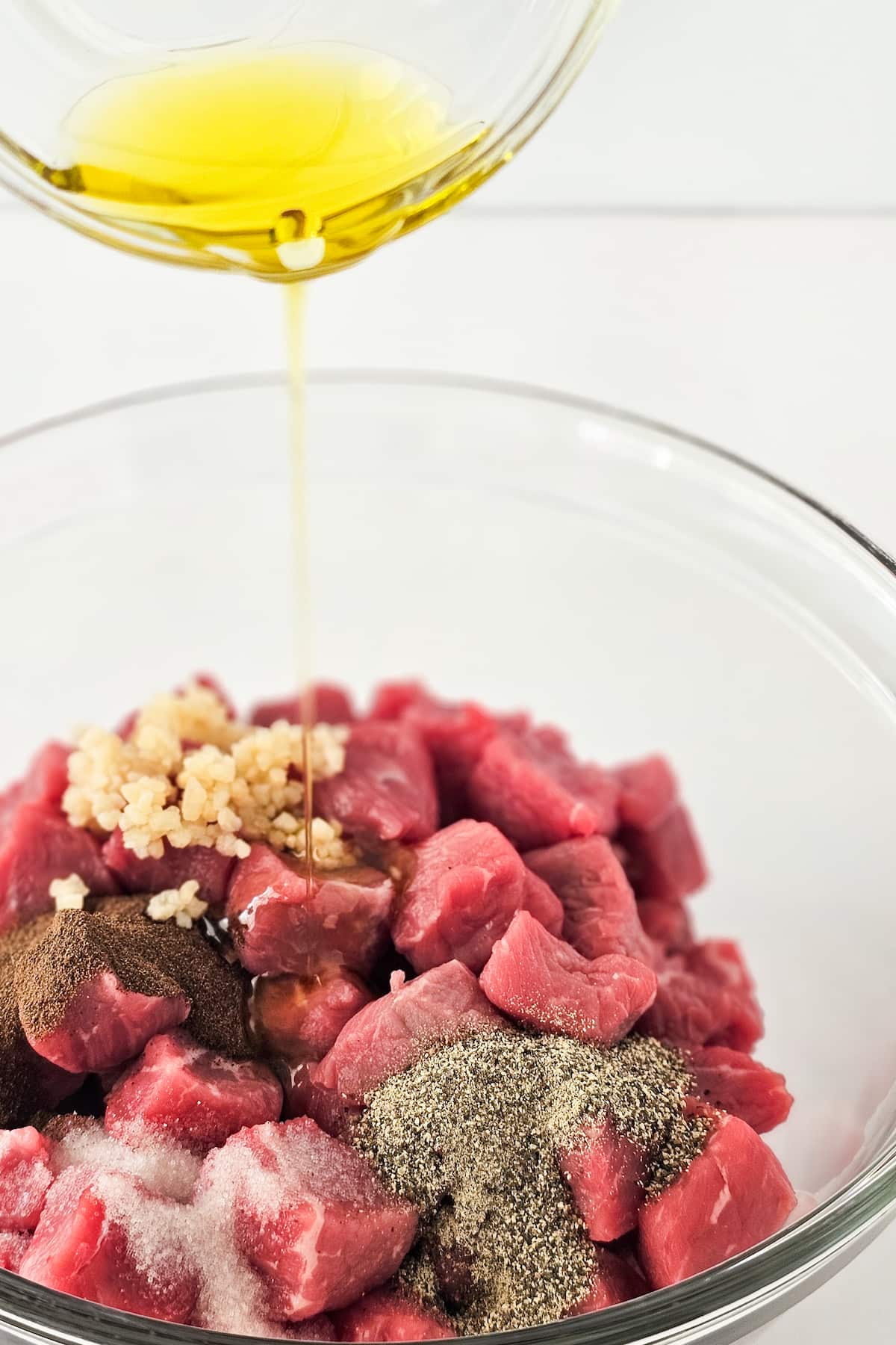 Chopped raw steak in a glass bowl with minced garlic, pepper, and spices, ready to become air fryer steak bites as olive oil is poured over it.