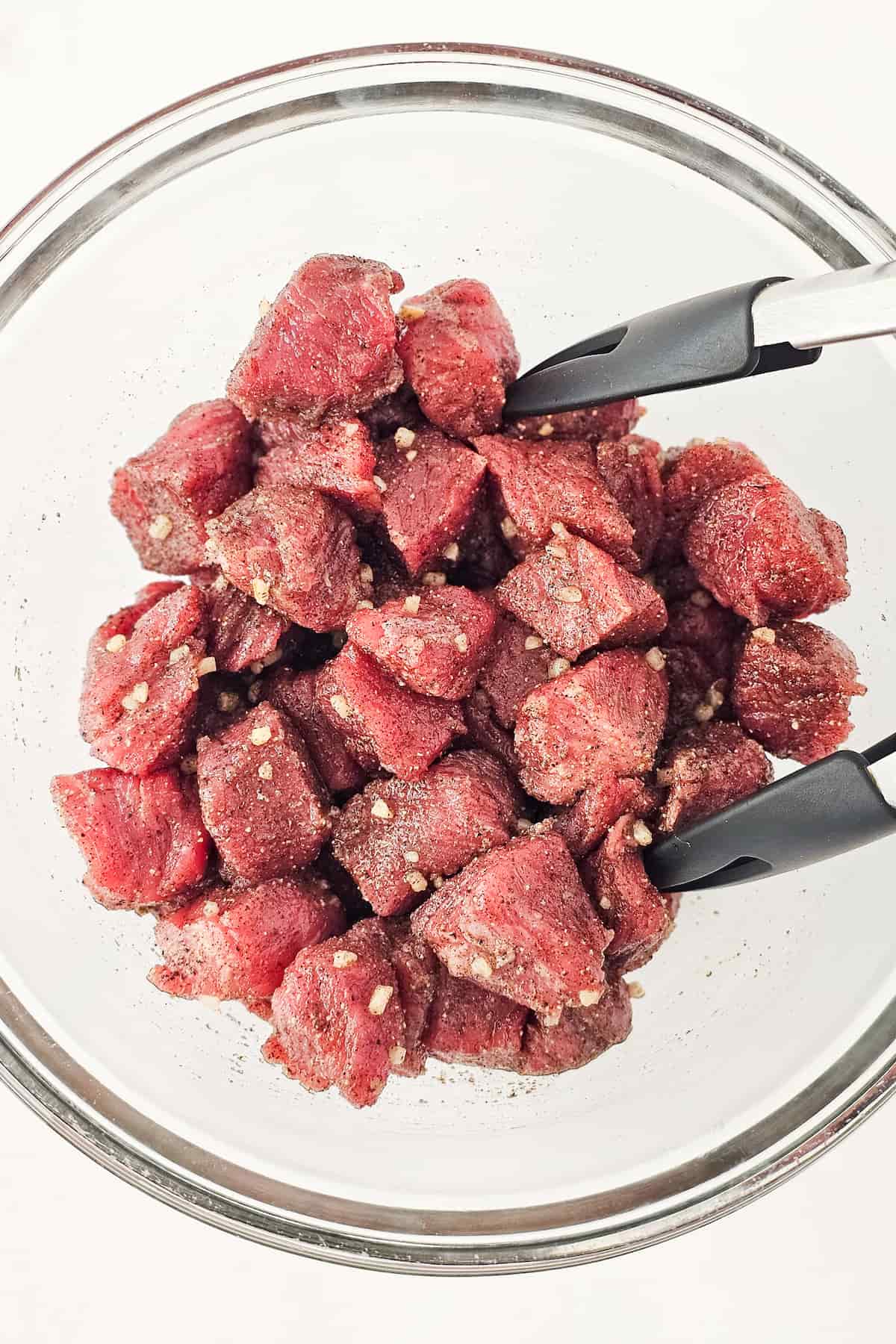 A glass bowl filled with raw steak pieces, seasoned with spices for mouthwatering air fryer steak bites, is being held by black-handled tongs.