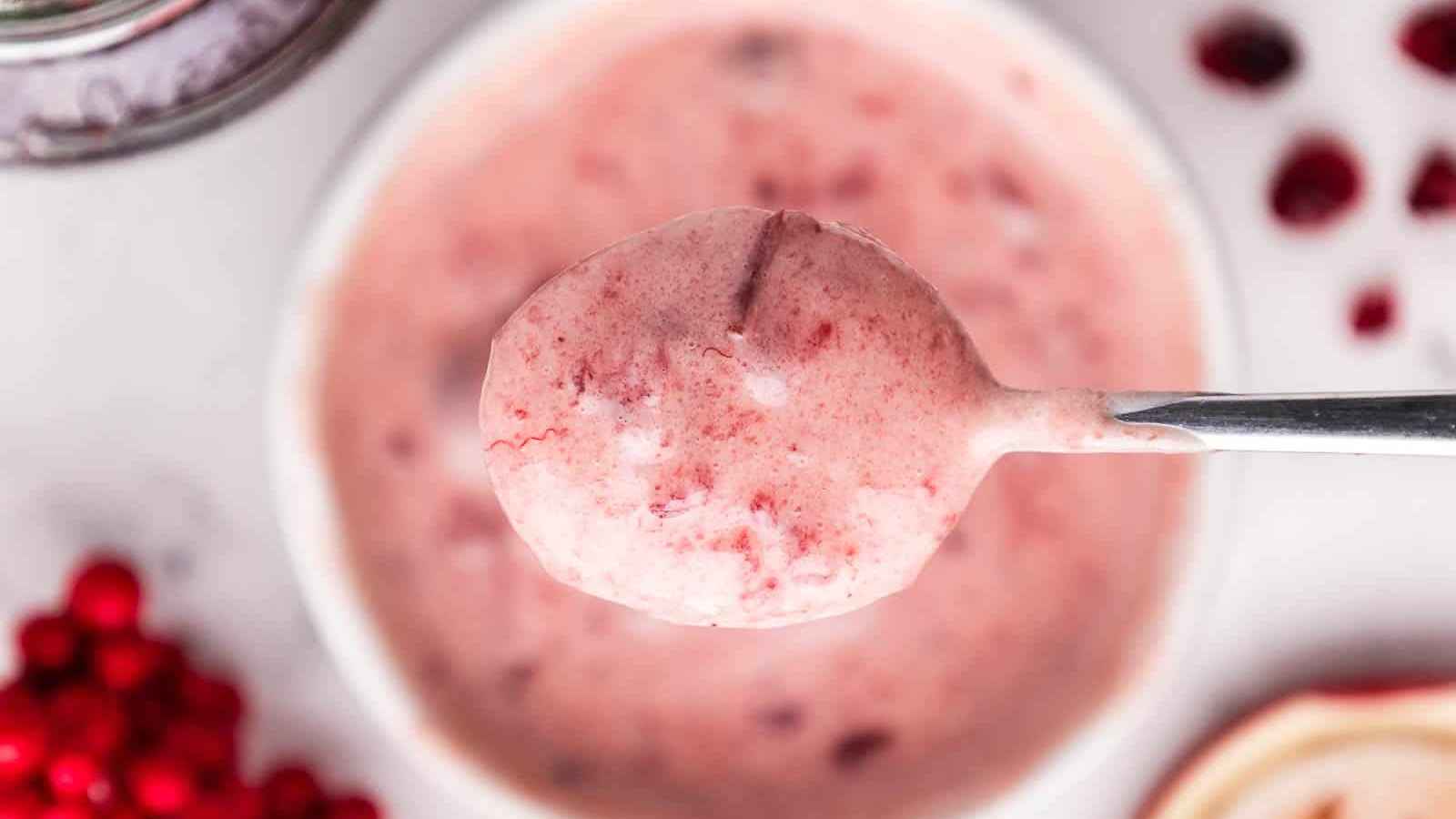 A spoonful of creamy pink dessert with red berry bits over a bowl, surrounded by berries and blurred elements.