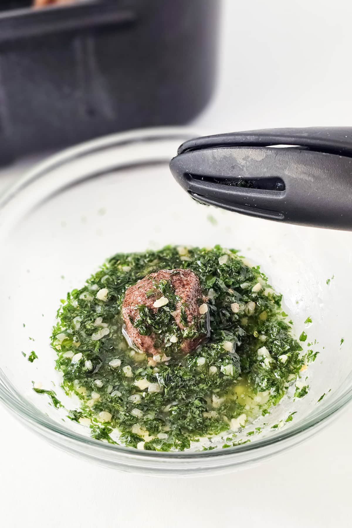 A piece of cooked steak, destined to become savory air fryer steak bites, is partially submerged in a bowl of melted butter, parsley, and minced garlic. A pair of black tongs hovers above the bowl, ready to perfect the bite-sized delicacies.