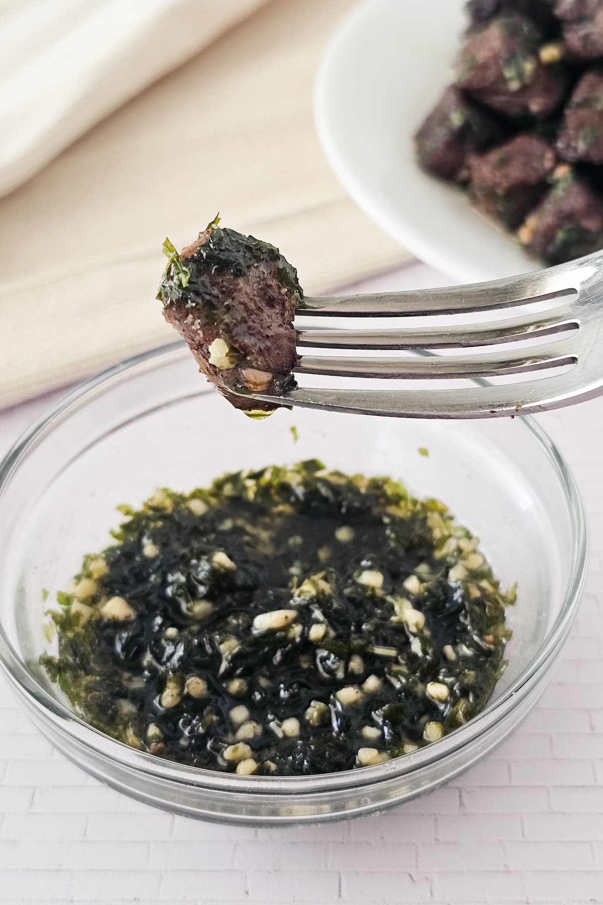 A fork holds a piece of steak above a bowl of garlic butter with parsley. More air fryer steak bites are on a plate in the background.