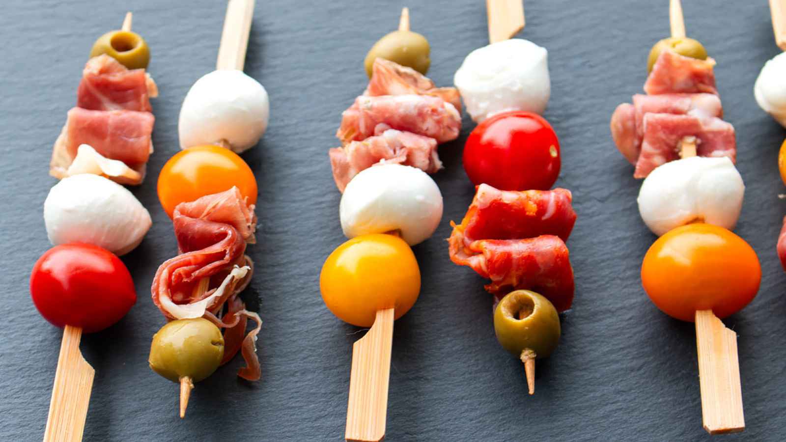 Skewers with cherry tomatoes, mozzarella balls, cured meat, and olives on a dark surface.