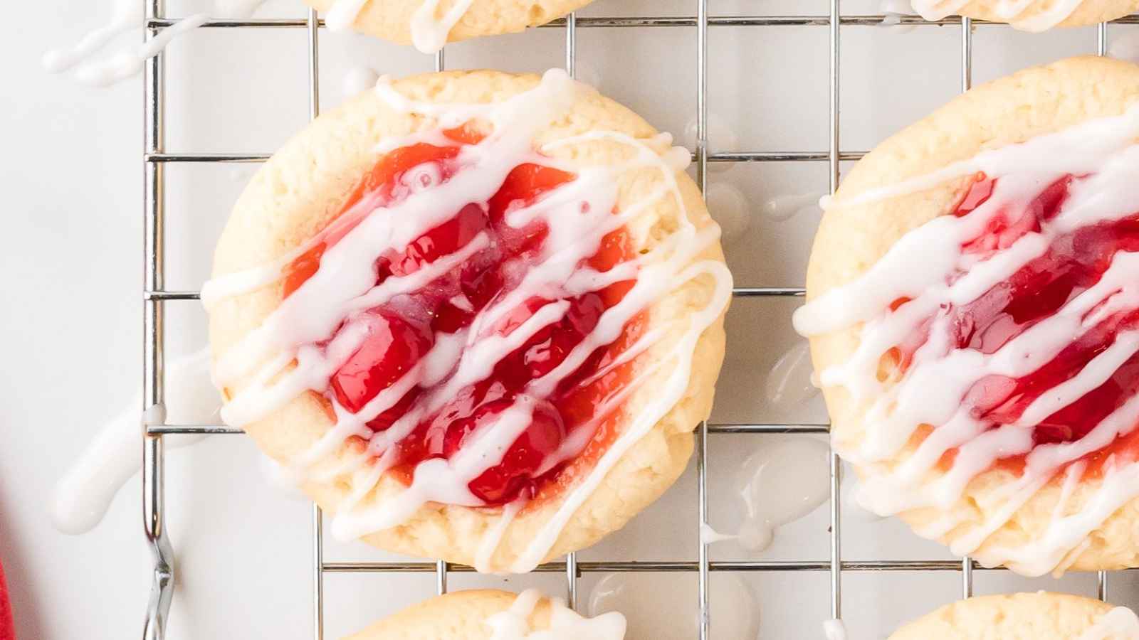 Close-up of cookies topped with red jam and white icing, placed on a wire cooling rack.