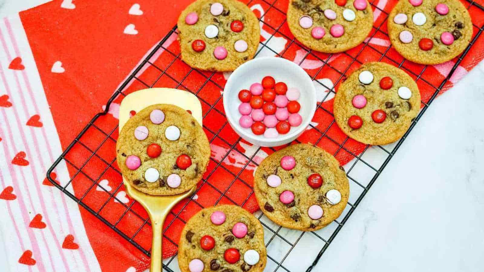 A cooling rack with chocolate chip cookies topped with red, pink, and white candies. A small bowl of candies is on the rack. A red cloth with white hearts is underneath.