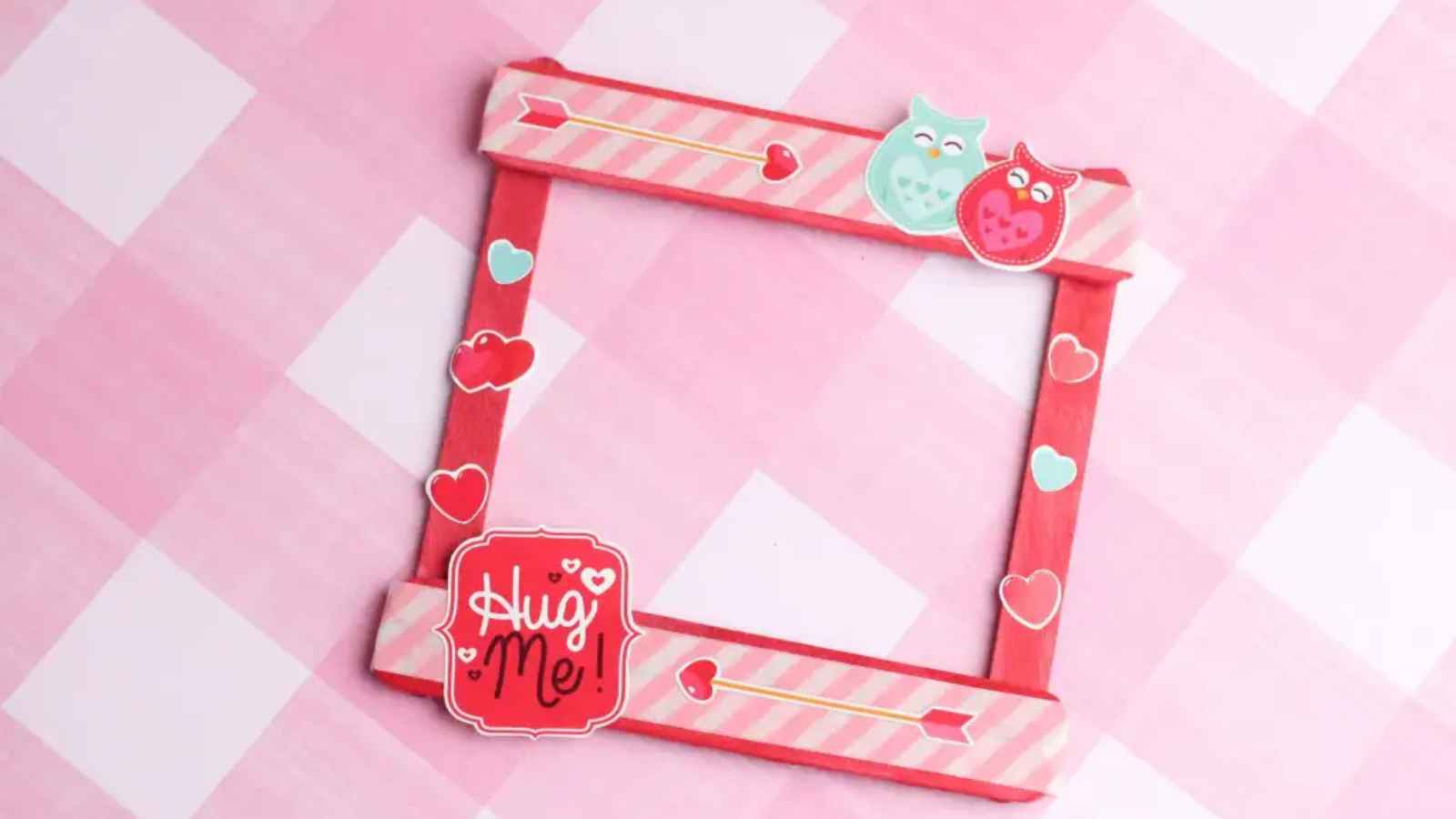 A pink and red craft frame with heart designs, featuring two cartoon owls and a "Hug Me!" sign at the bottom left corner, on a pink checkered background.