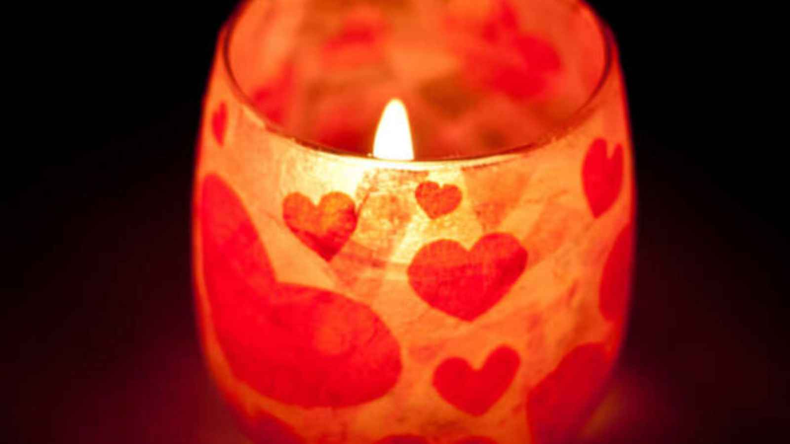 A lit candle in a glass holder with red heart patterns, creating a warm glow against a dark background.