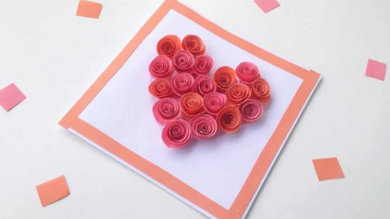 Handmade card with paper roses arranged in a heart shape on a white square with an orange border, surrounded by scattered orange paper squares.