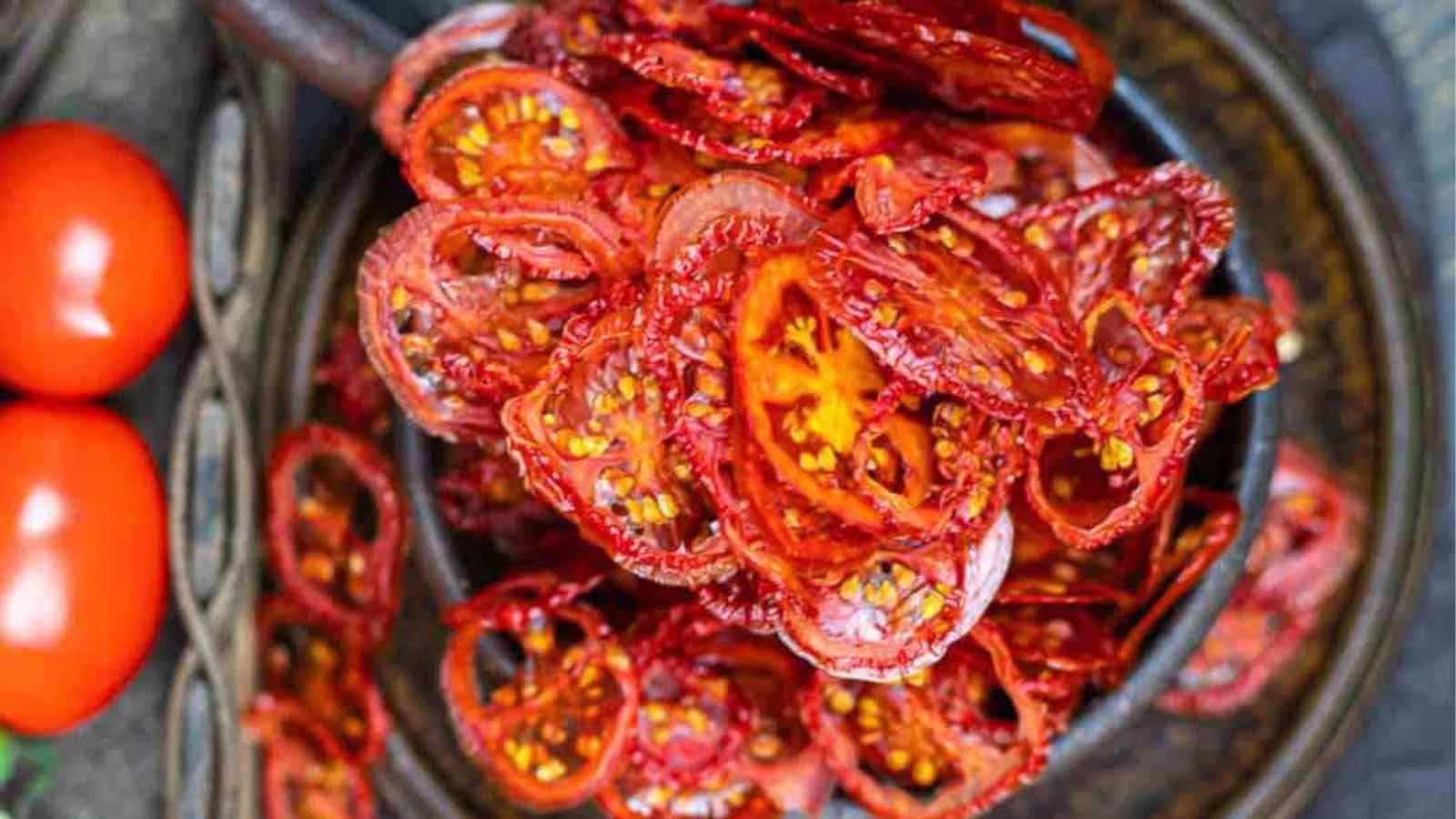 A basket of fresh whole tomatoes next to a plate of sliced, sun-dried tomatoes on a dark textured surface.