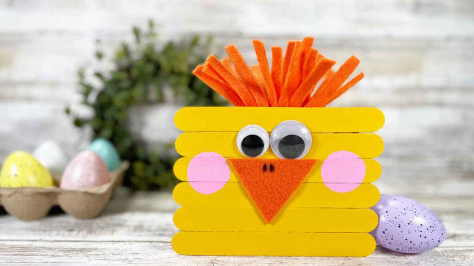 Crafted chick made from yellow popsicle sticks with googly eyes, orange felt beak, and orange hair against a wooden background.