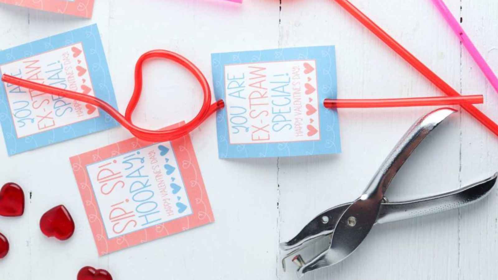 Colorful drinking straws with heart and straight shapes, attached to cards reading "You are ex-straw special!" and "Sip, sip, hooray!" A hole punch is nearby on a white surface.