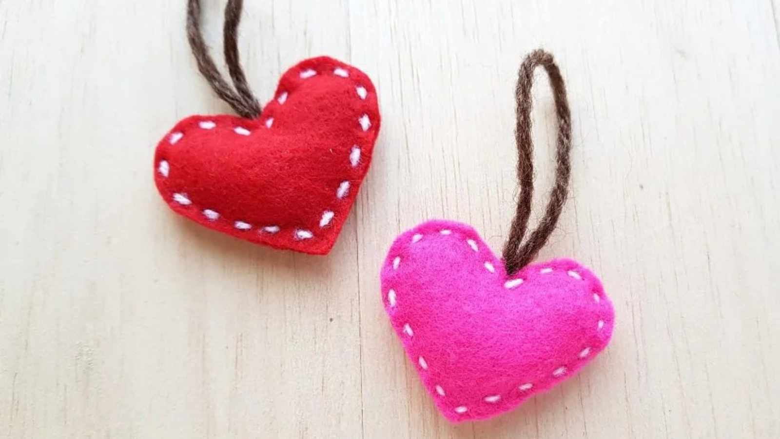 Two felt heart ornaments, one red and one pink, with white stitching and brown loops against a light wooden background.