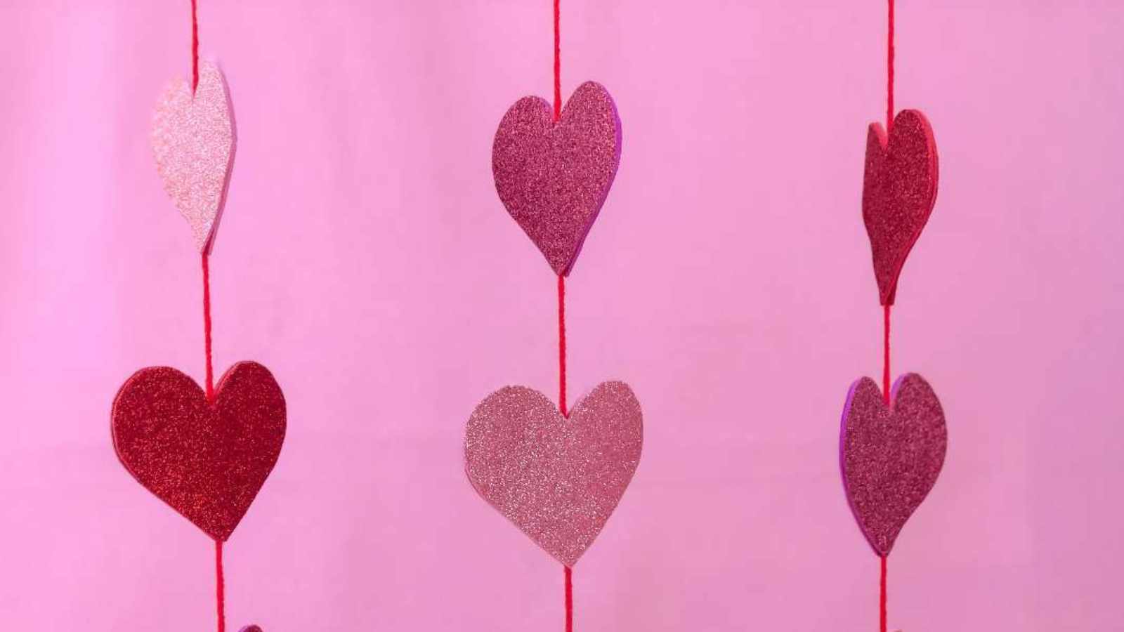 Several glittery heart cutouts hang vertically on red strings against a pink background.