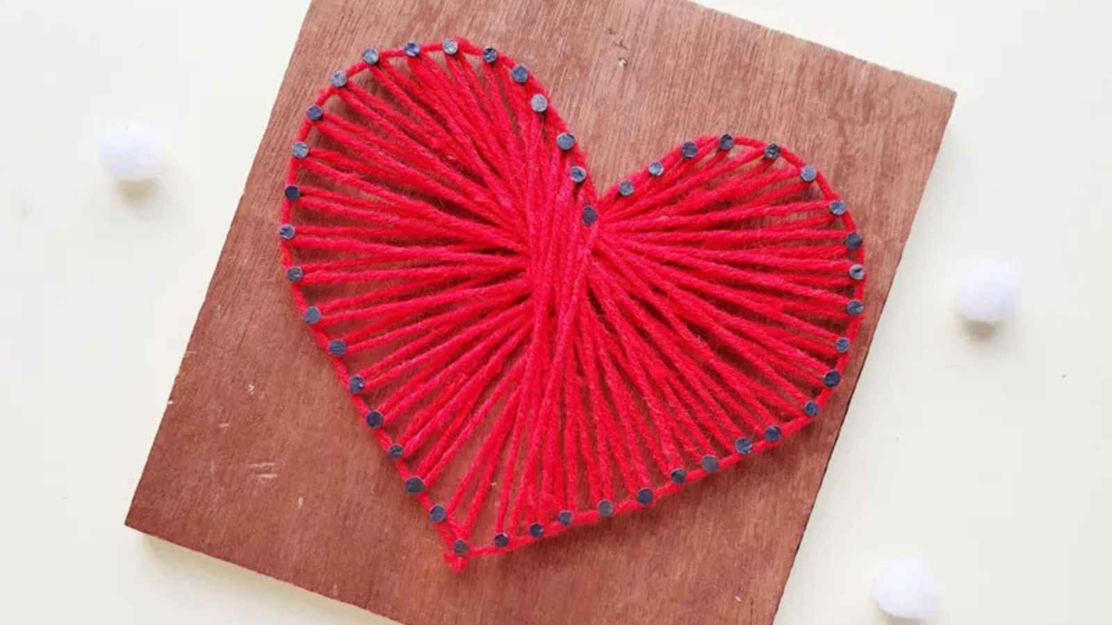 A red string heart design on a wooden board, with nails outlining the heart shape and the string threaded between them.