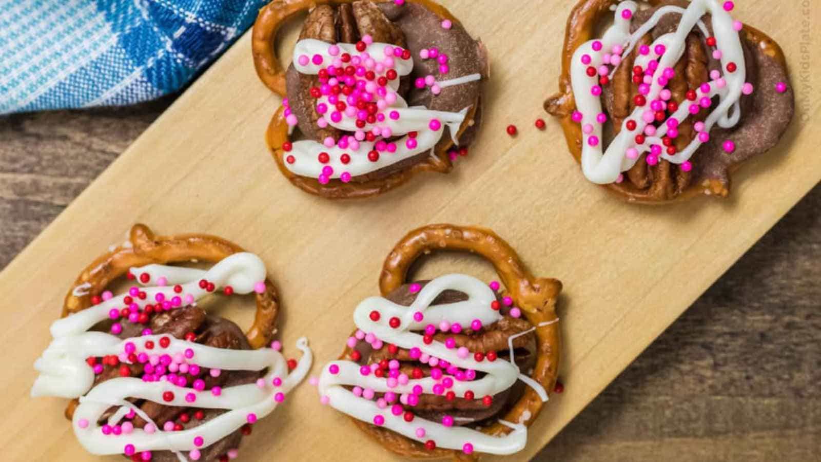 Four pretzel snacks topped with chocolate, white icing, and pink sprinkles on a wooden board; blue checkered cloth partially visible.