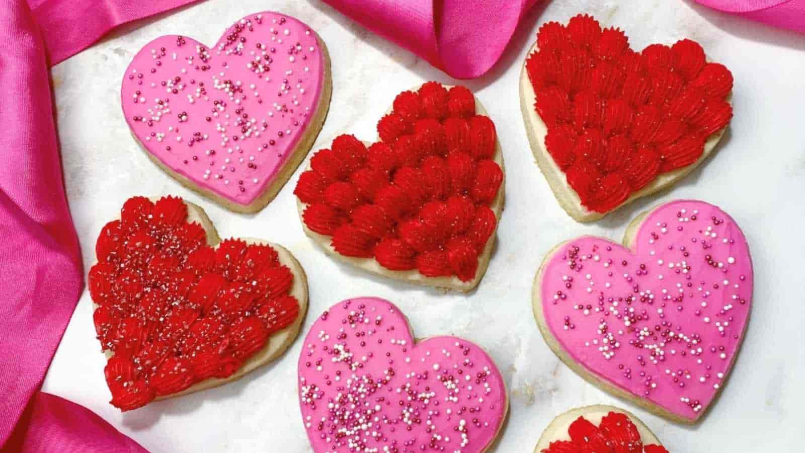 Heart-shaped cookies with pink and red icing, topped with silver and pink sprinkles, arranged on a white surface with pink fabric.