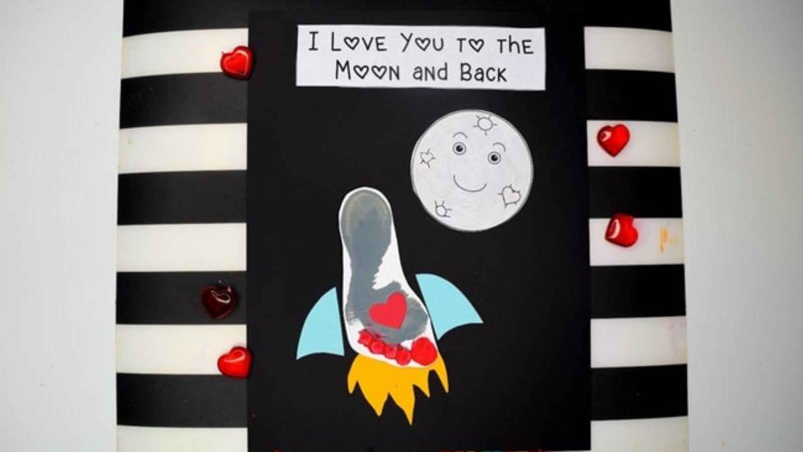 A craft with a foot-shaped rocket on a black background, a gray moon with a smiley face, and the text "I love you to the moon and back." Red heart decorations are on the sides.