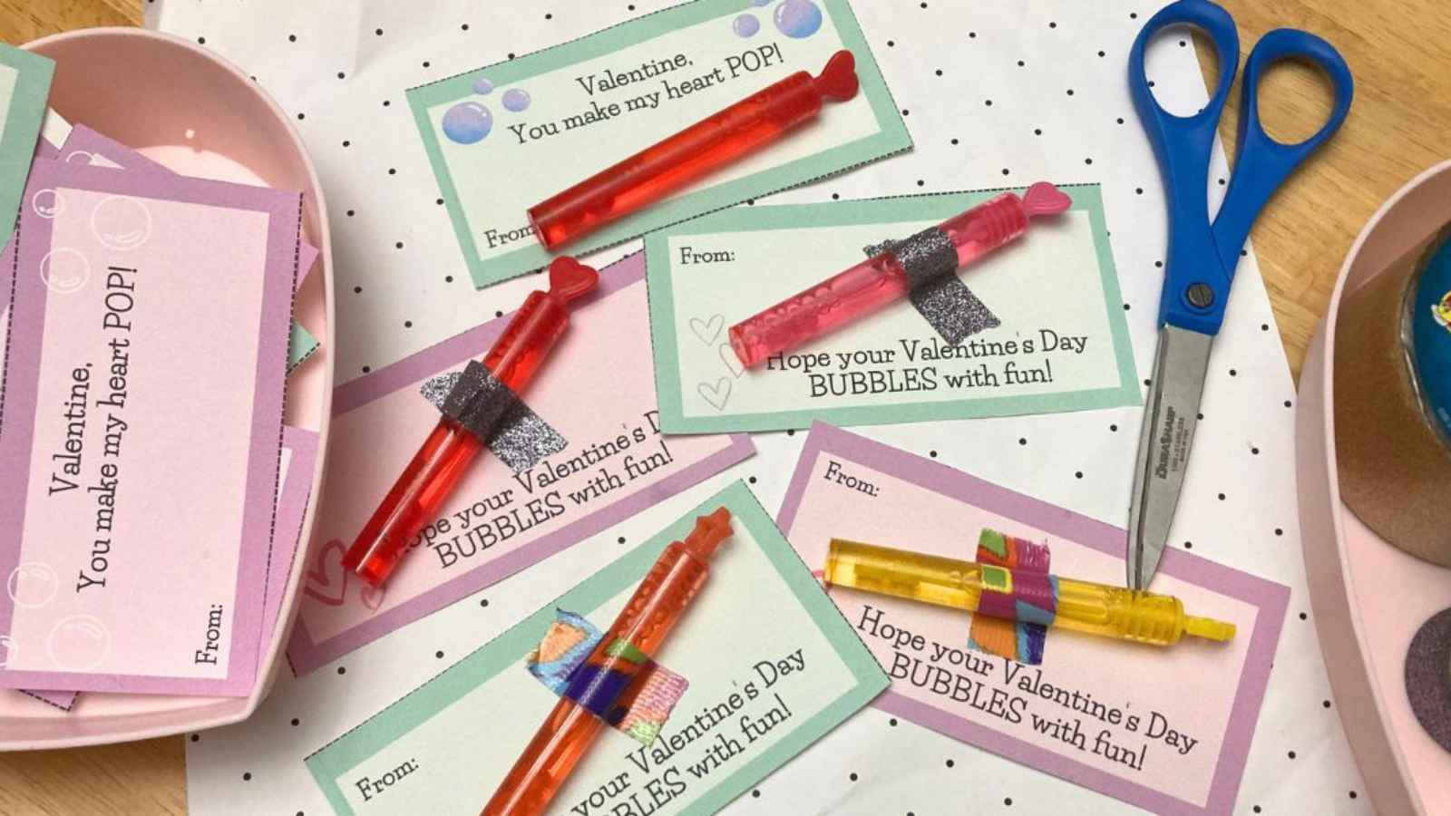 Handmade Valentine's cards with bubble wands, scissors, and tape on a polka dot surface. Cards read phrases like "You make my heart POP!" and "BUBBLES with fun!.
