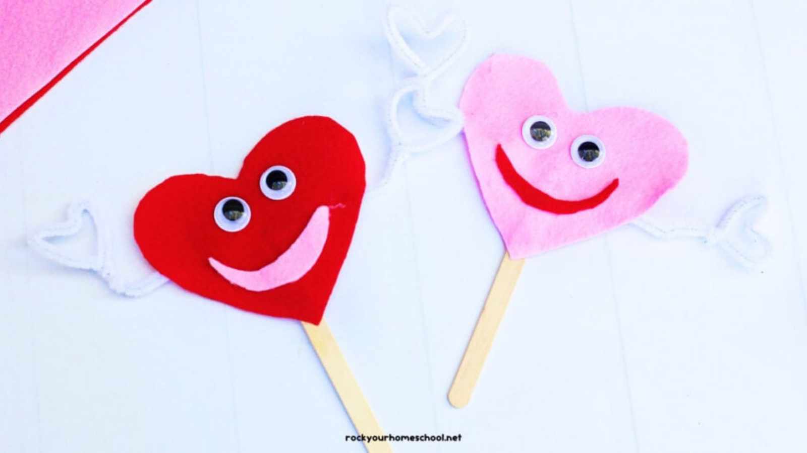 Two felt heart crafts with googly eyes and smiles on sticks, one red and one pink, with white pipe cleaner arms.