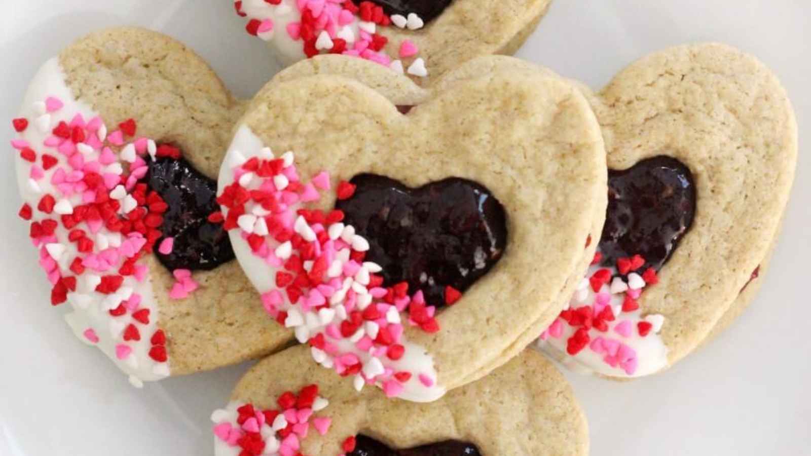Heart-shaped cookies with jam centers, white icing, and pink and red sprinkles on a plate.