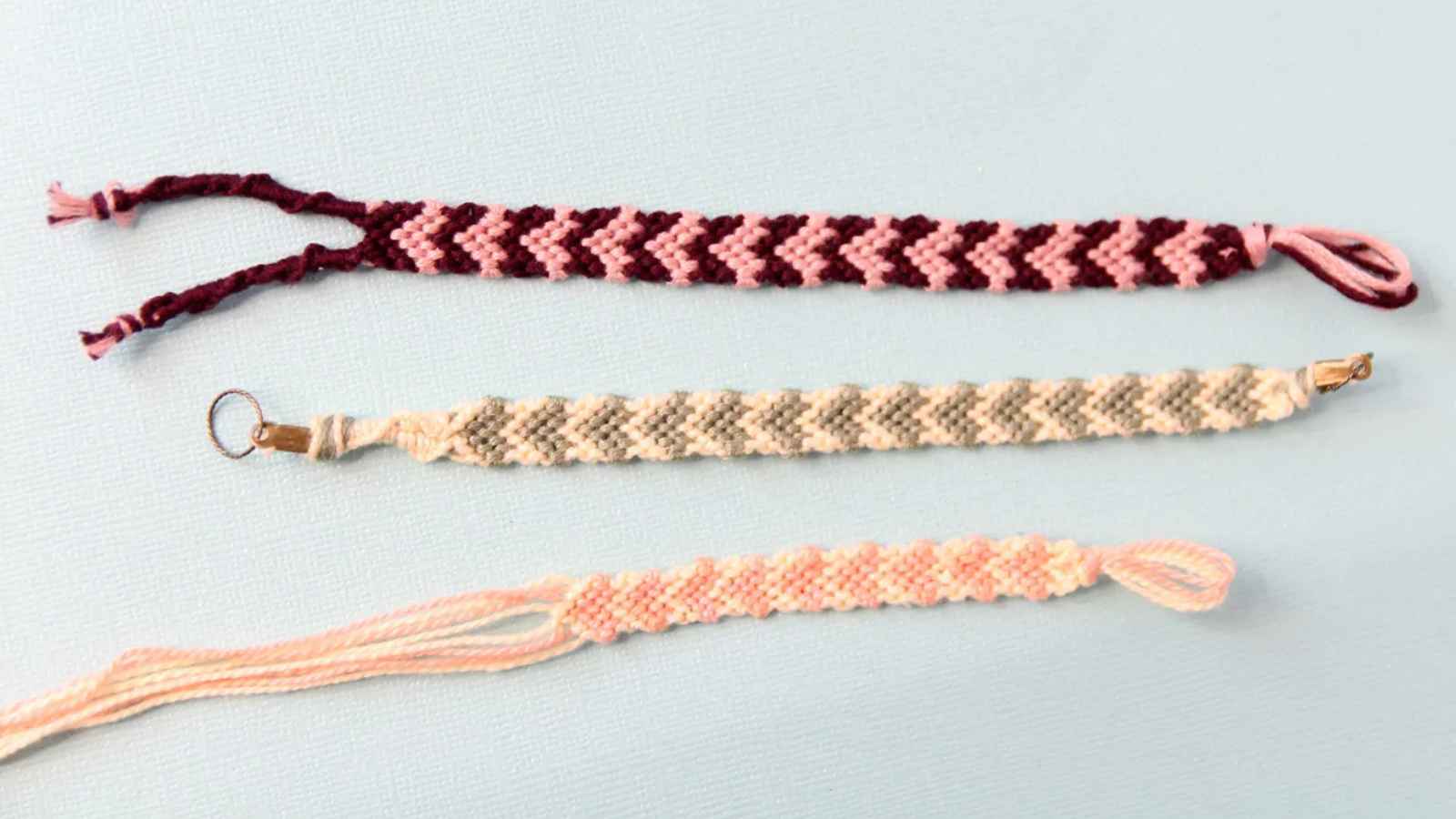 Three woven friendship bracelets on a light blue background, featuring chevron and diamond patterns in shades of pink, maroon, and beige.