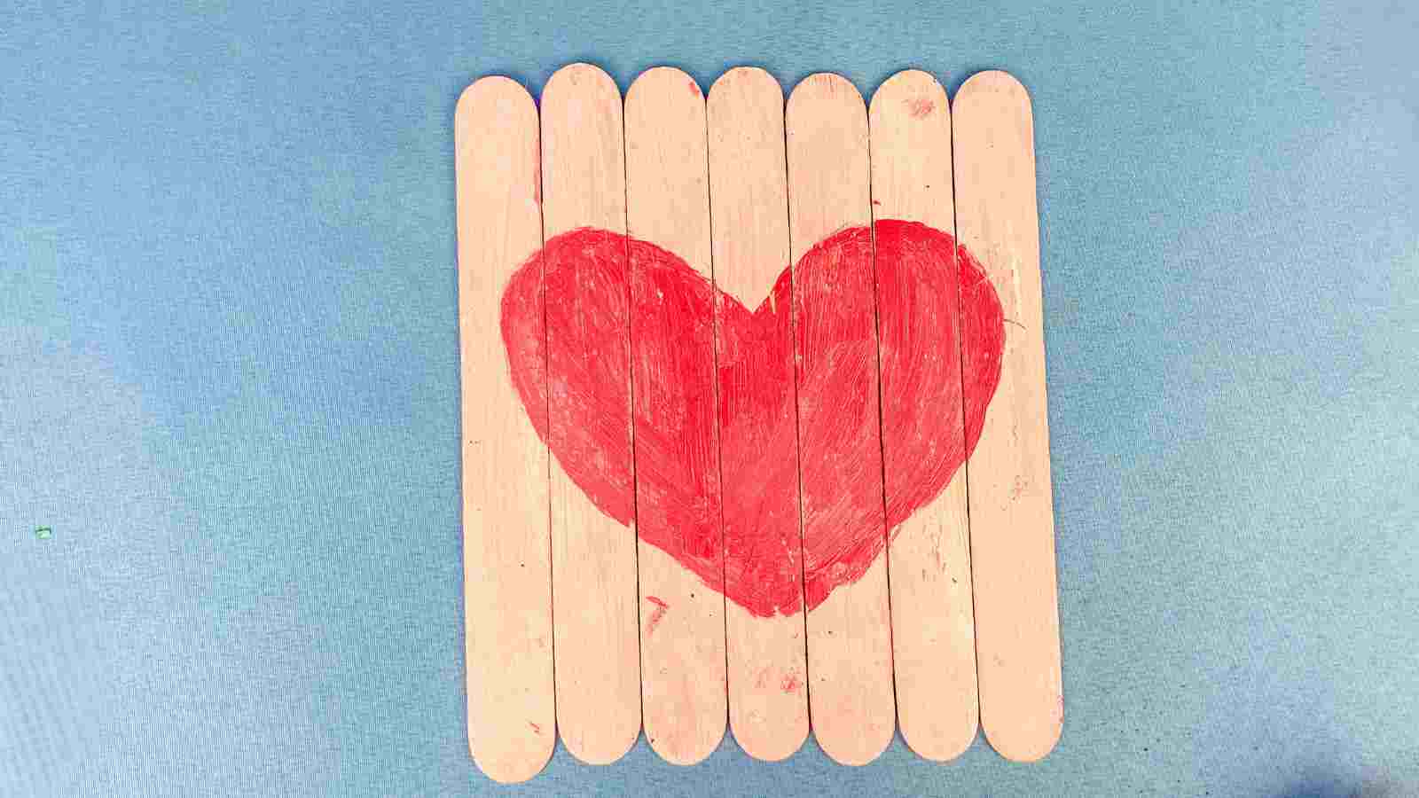 Craft sticks arranged in a square with a red heart painted on them, set against a blue background.