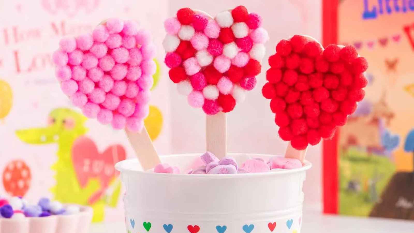 Three heart-shaped crafts made of colored pom-poms on sticks are placed in a white cup decorated with hearts.