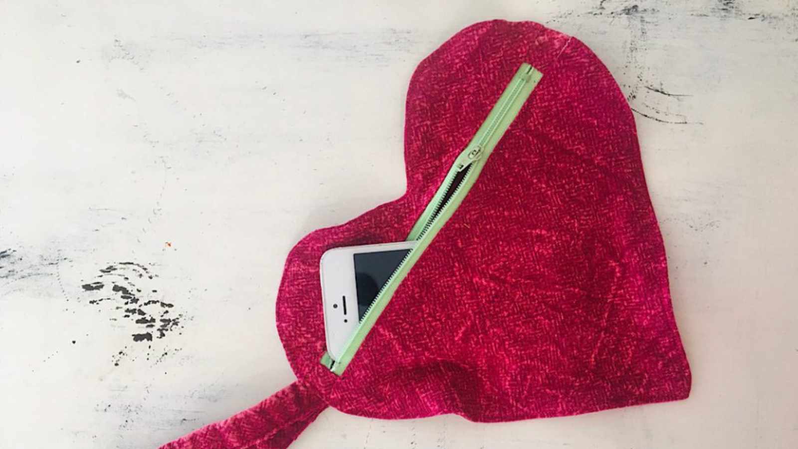 Heart-shaped pink pouch with a green zipper partially open to reveal a smartphone inside, placed on a white surface.