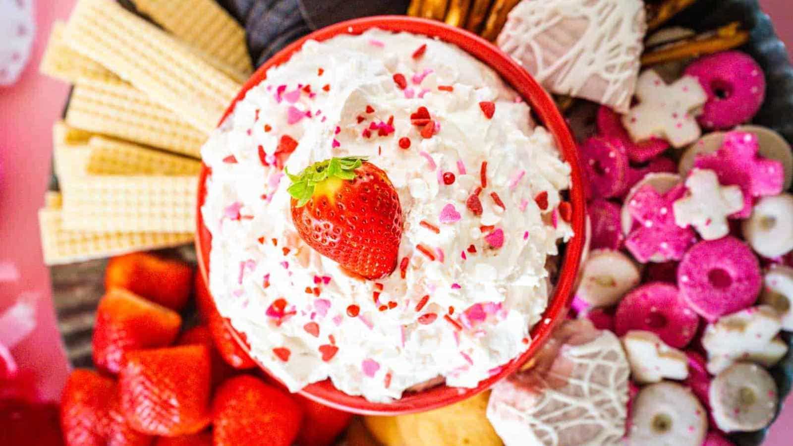 A bowl of whipped cream topped with a strawberry sits in the center, surrounded by wafers, strawberries, pink and white cookies, and chocolate-covered treats on a platter.