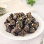 A plate of air fryer steak bites, garnished with herbs and garlic, rests on a light-colored surface. A fork and a small bowl of sauce linger in the background, tempting the senses with their inviting presence.
