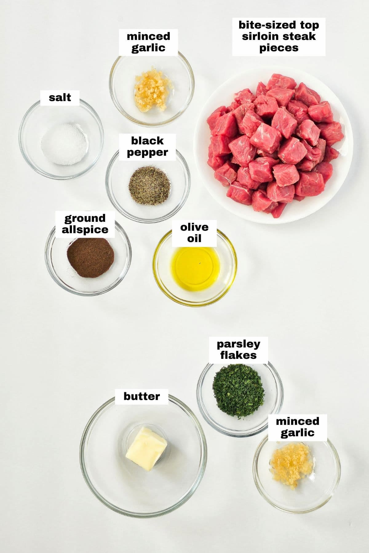 Ingredients for a recipe: air fryer steak bites made with sirloin steak pieces, minced garlic, salt, black pepper, ground allspice, olive oil, butter, and parsley flakes—all labeled and neatly placed in bowls.