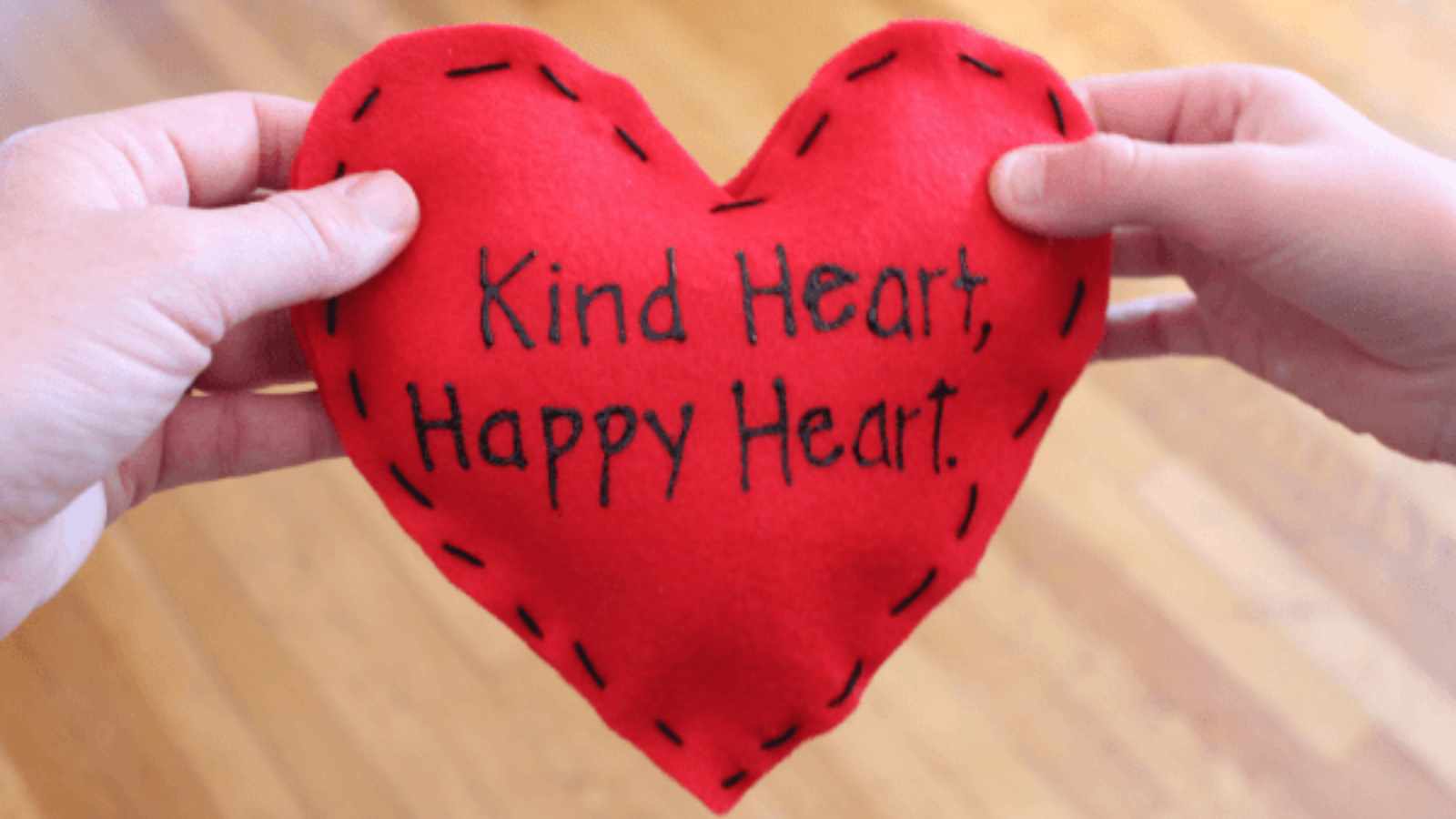 Two hands hold a red heart-shaped fabric with black stitching and the words "Kind Heart, Happy Heart.