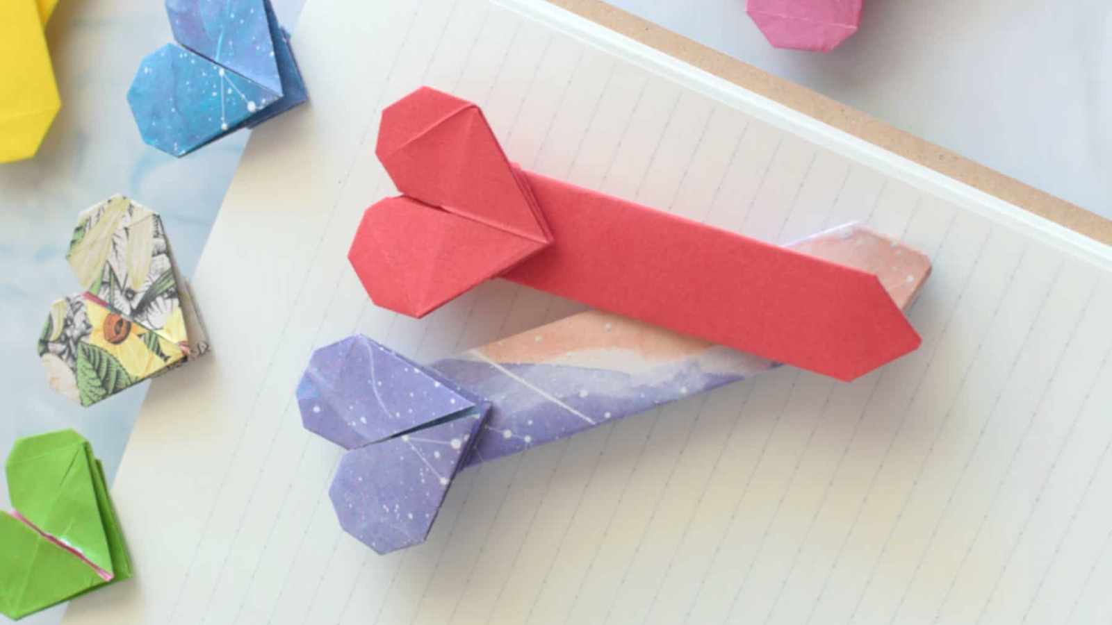 Colorful origami bookmarks shaped like arrows with heart-shaped ends rest on an open notebook with lined pages.
