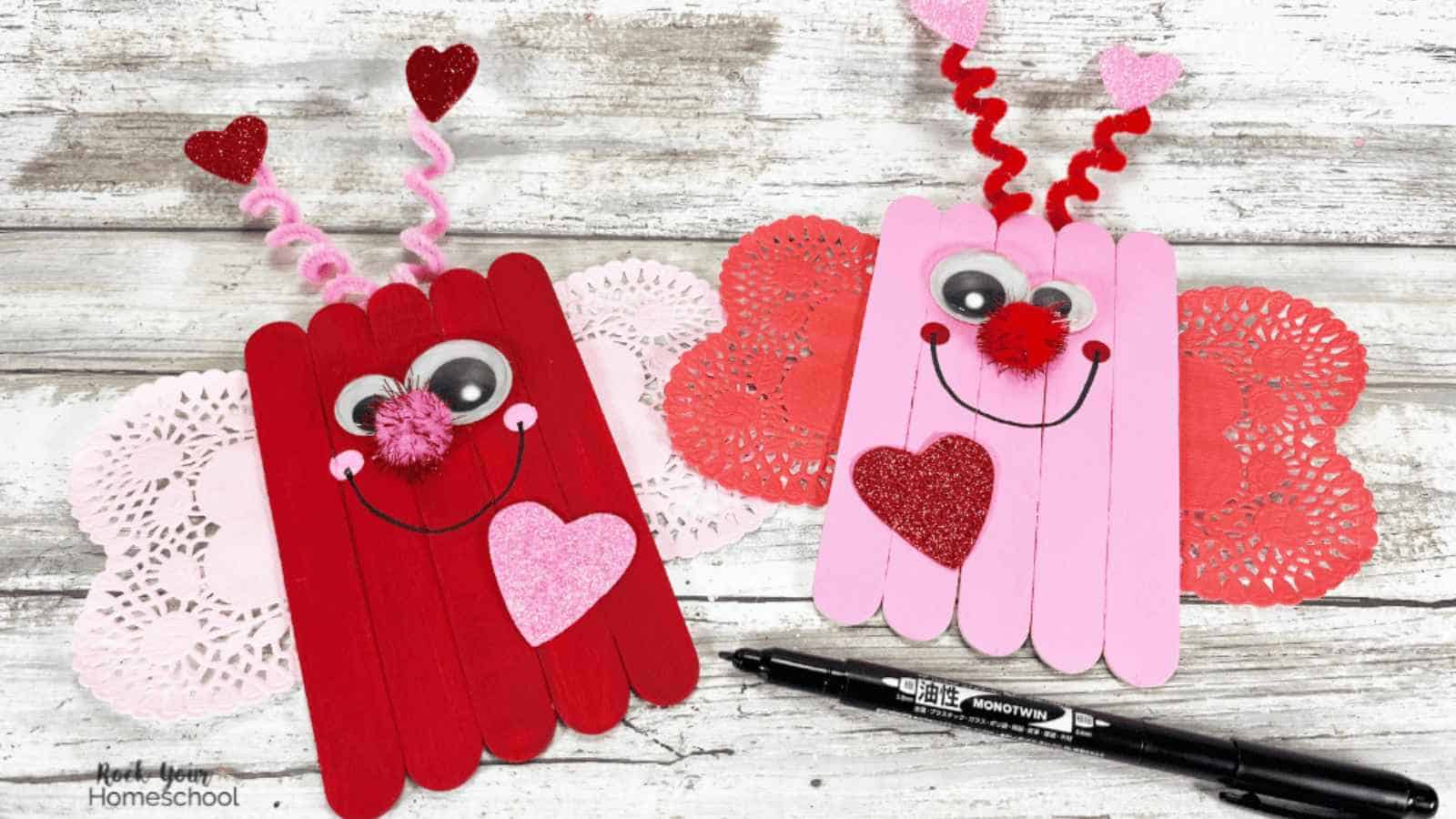 Two butterfly crafts made from red and pink popsicle sticks, decorated with googly eyes, pompoms, and heart shapes, on a wooden surface with a black marker nearby.