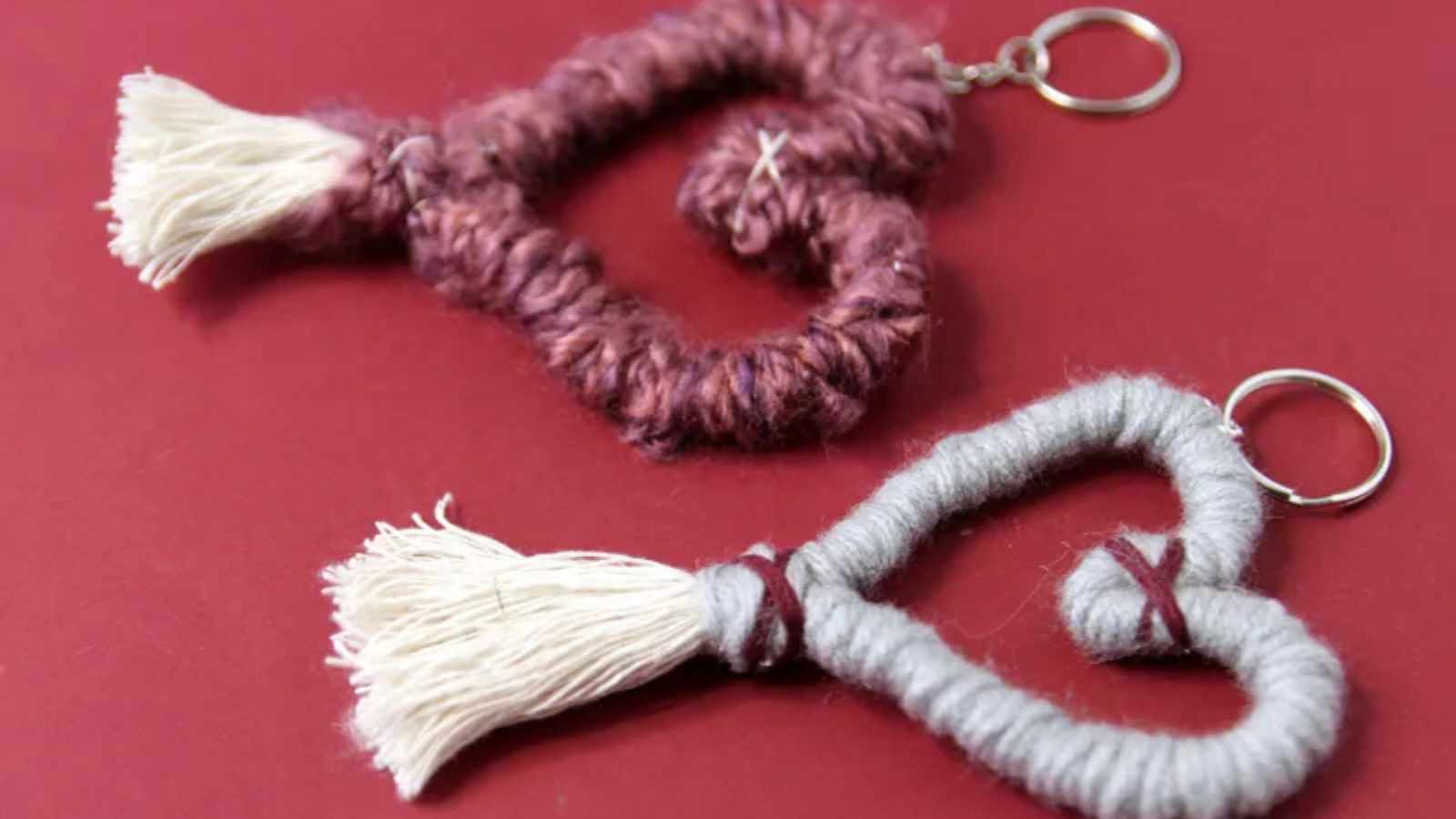 Two heart-shaped keychains made of yarn on a red surface. One is burgundy with a white tassel, and the other is gray with a white tassel.