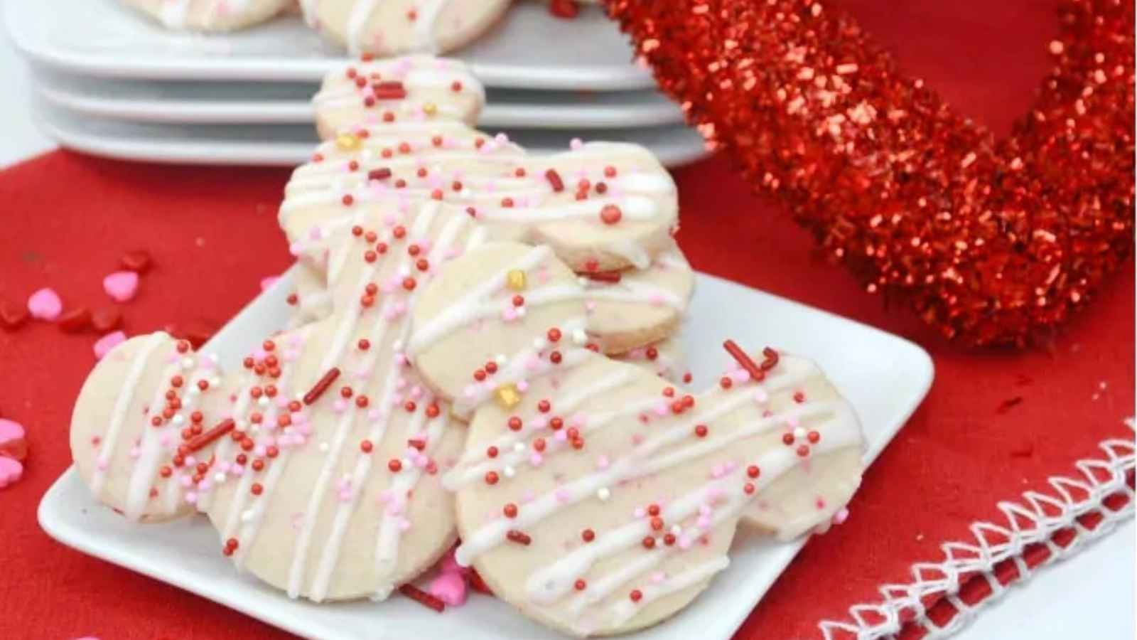 Mickey Mouse heart-shaped cookies with white icing and red, pink, and white sprinkles on a white plate. A red glittery garland decorates the background.