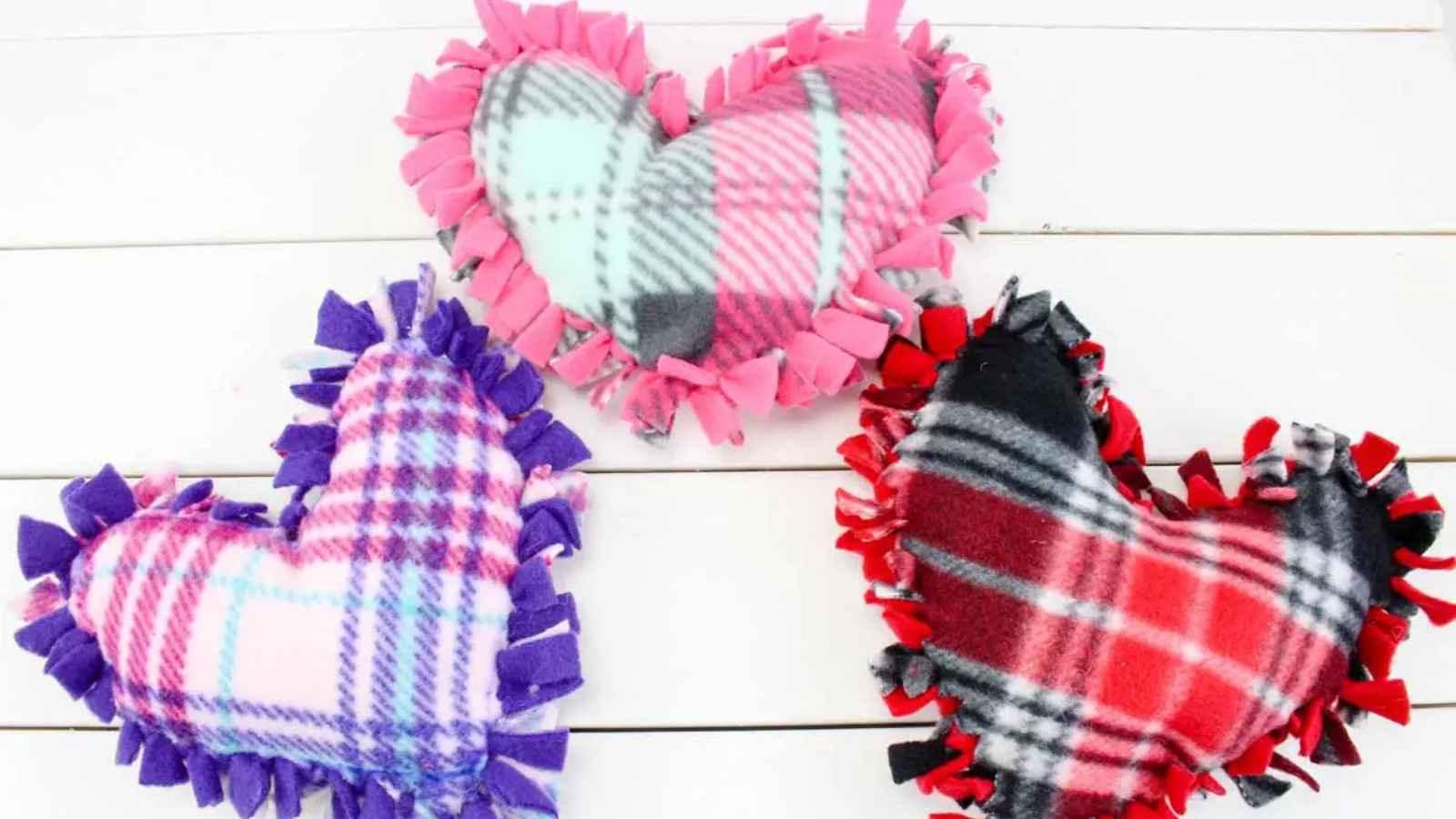 Three plaid heart-shaped pillows with fringed edges in pink, purple, and red color schemes, placed on a white wooden surface.