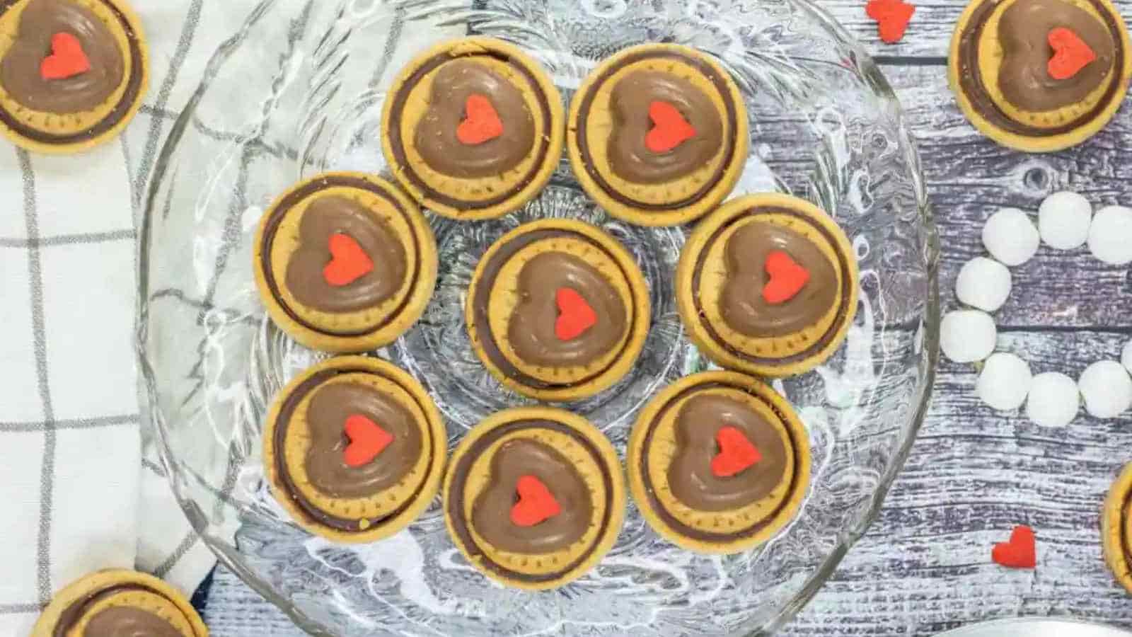 Glass plate with cookies topped with chocolate and red heart-shaped decorations on a wooden table.
