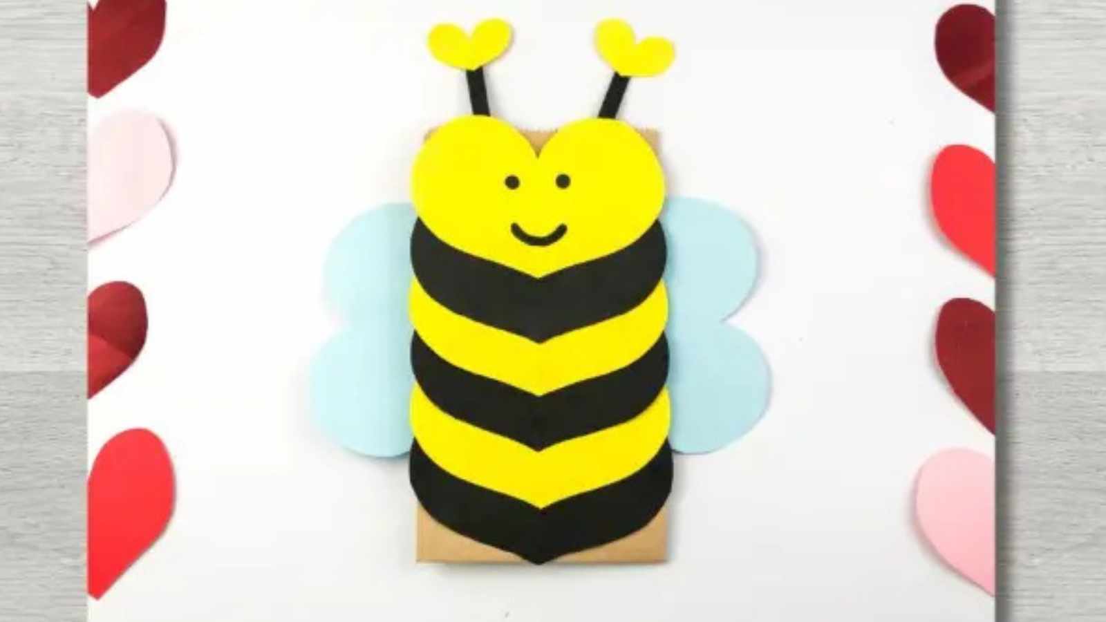 Crafted paper bee with heart-shaped wings and antennas, surrounded by red and pink paper hearts on a wooden surface.