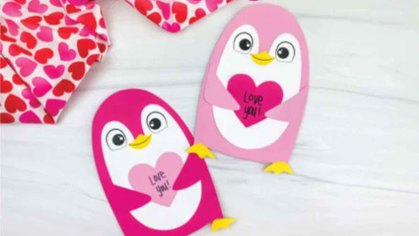Two pink penguin-shaped cards with "Love you!" written on their hearts are displayed on a light surface.