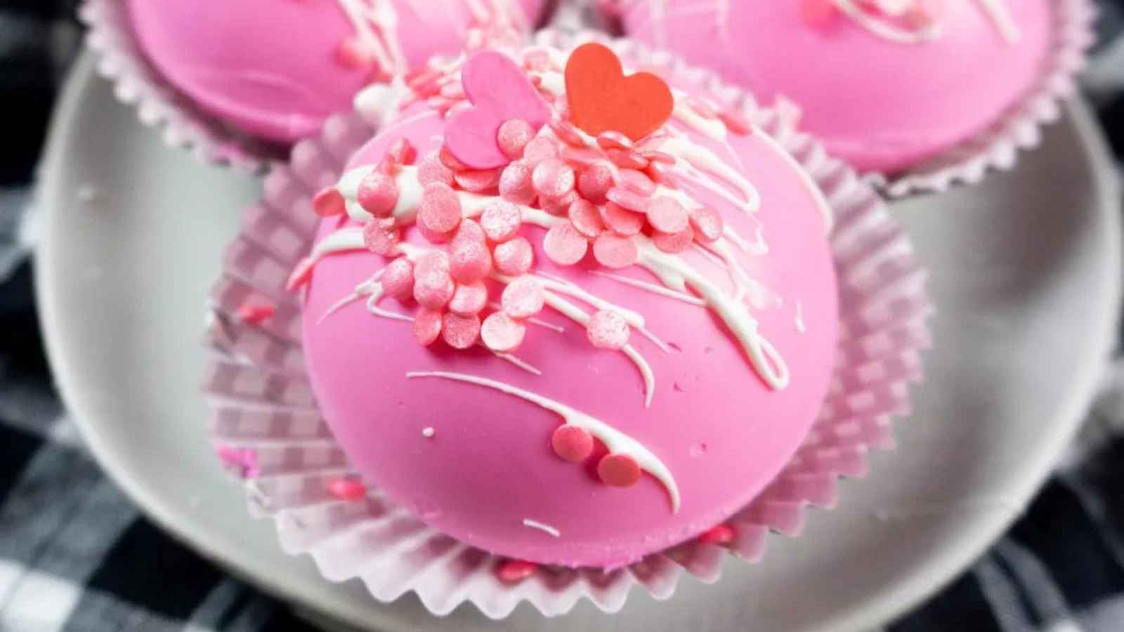 Pink Hot Cocoa Bombs with white drizzle and heart-shaped sprinkles arranged on a gray plate.