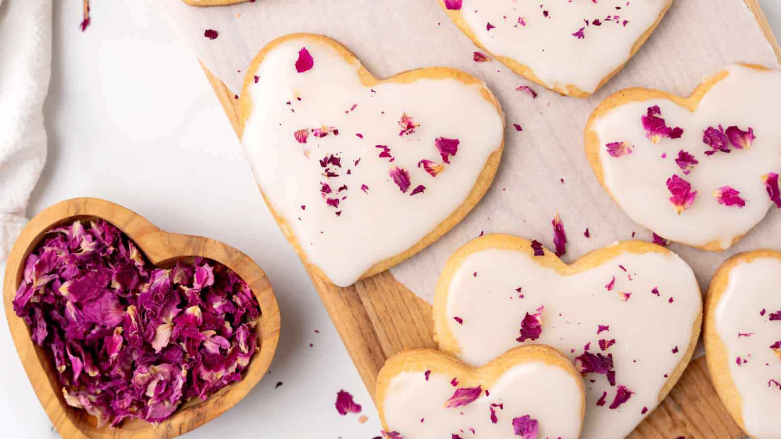 Heart-shaped cookies with white icing and rose petals on a wooden board, next to a heart-shaped bowl filled with dried rose petals.